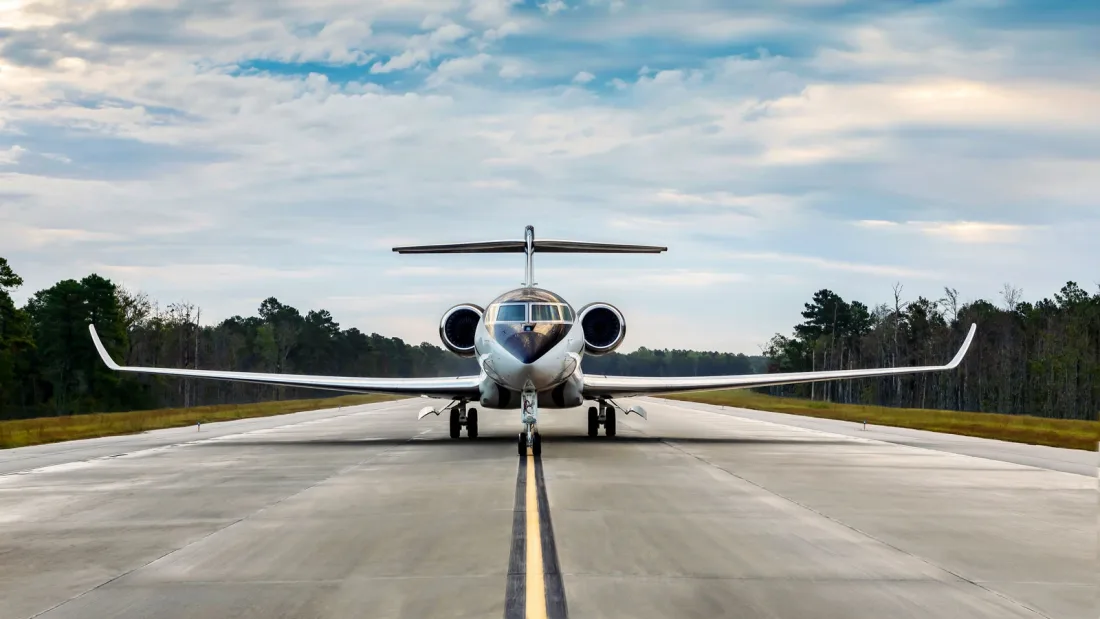 A white Gulfstream G700 business jet seen head-on on a runway centreline, with forest on both sides under a cloudy sky.
