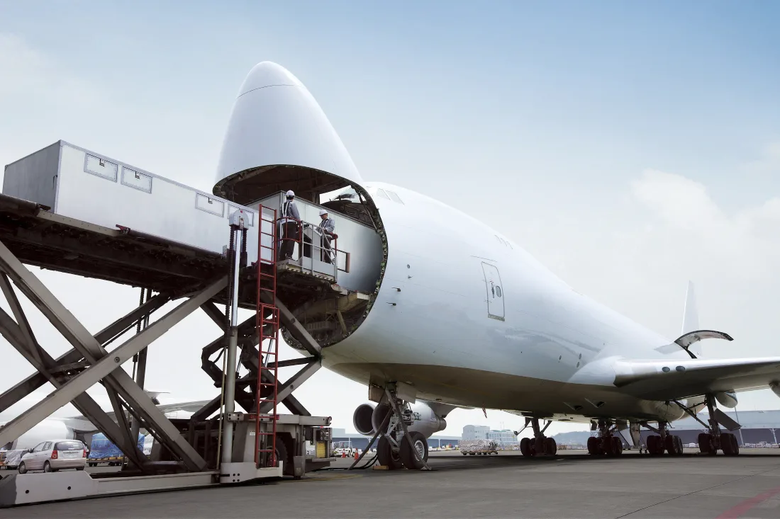 A Boeing 747 freighter with its nose door raised, two workers on a high loader platform at the opening.